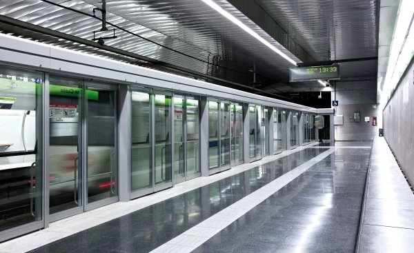 Andén de una estación de metro moderna con puertas de mampara automáticas de cristal y un tren en movimiento visible a través de ellas.