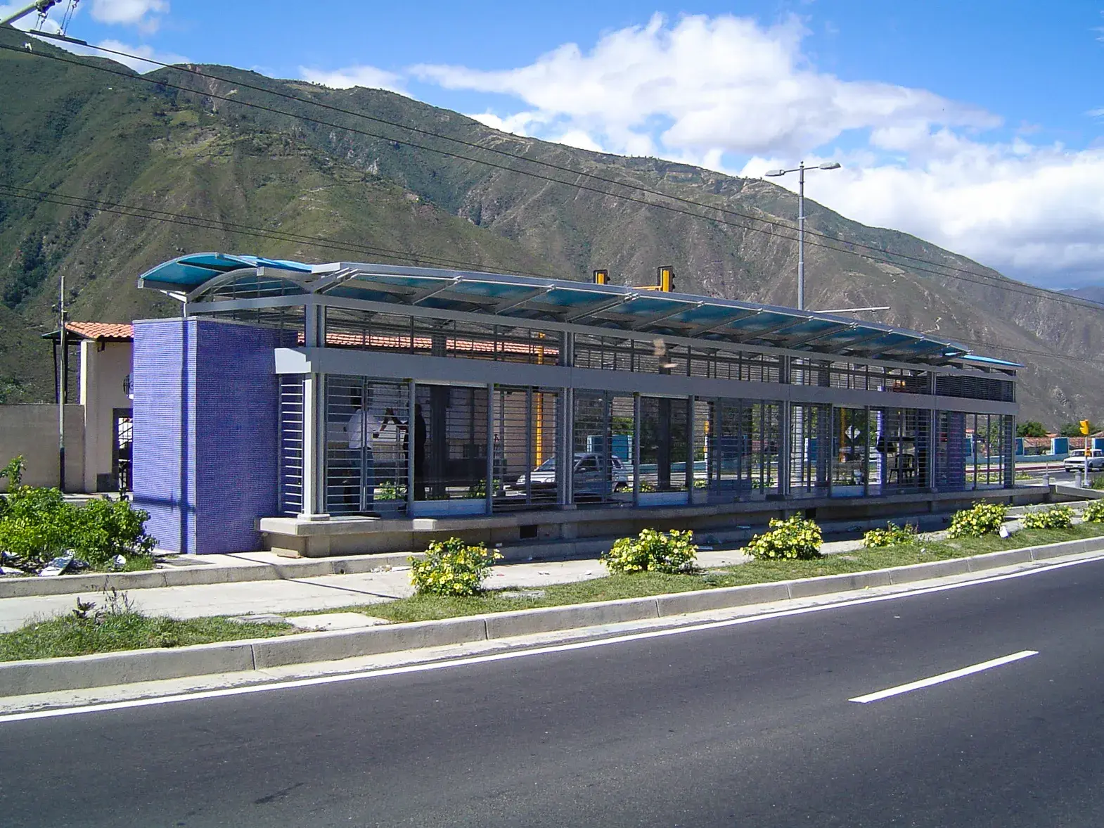 Estación de autobús moderna con estructura metálica y paredes de color morado, situada junto a una carretera con montañas al fondo.