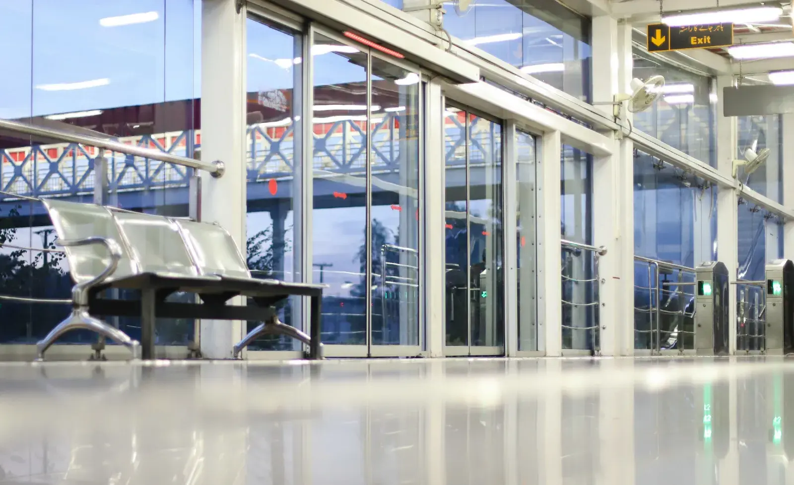 Vista a nivel de suelo de una estación de transporte, mostrando el reflejo del interior, bancos metálicos y puertas automáticas de cristal.