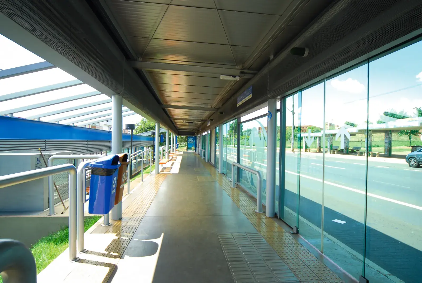 Pasillo interior de una estación de BRT con suelo podotáctil, paredes de cristal y papeleras de reciclaje, con vista a la vía del autobús.