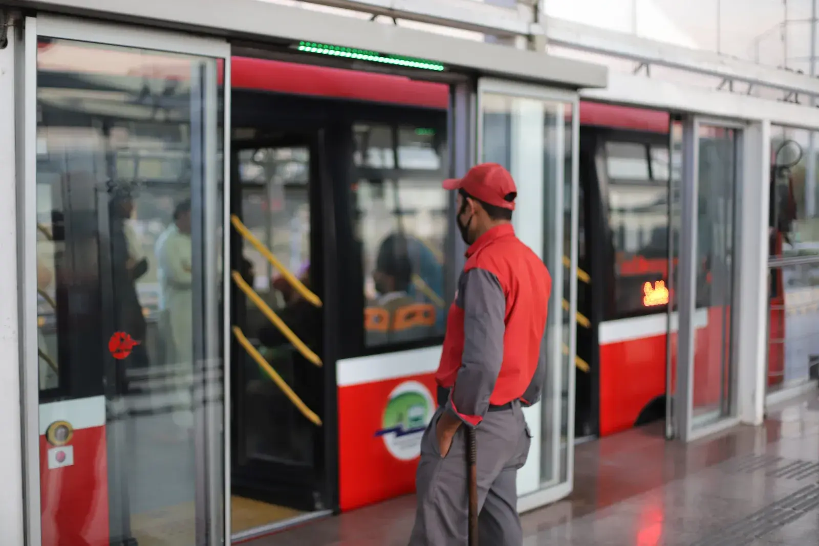 Un miembro del personal de la estación, con gorra roja y mascarilla, de pie junto a las puertas corredizas de vidrio de una estación de BRT, mientras un autobús rojo y blanco está parado en el andén.