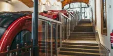 Interior de una estación con un funicular rojo moderno y puertas automáticas de vidrio a lo largo del andén con escaleras lateral.