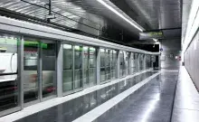 Andén de una estación de metro moderna con puertas de mampara automáticas de cristal y un tren en movimiento visible a través de ellas.