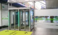 Pasillo unidireccional antirretorno con puertas automáticas de cristal y luces LED verdes, señalizado con un cartel amarillo de 'Saída/Exit' en el suelo.