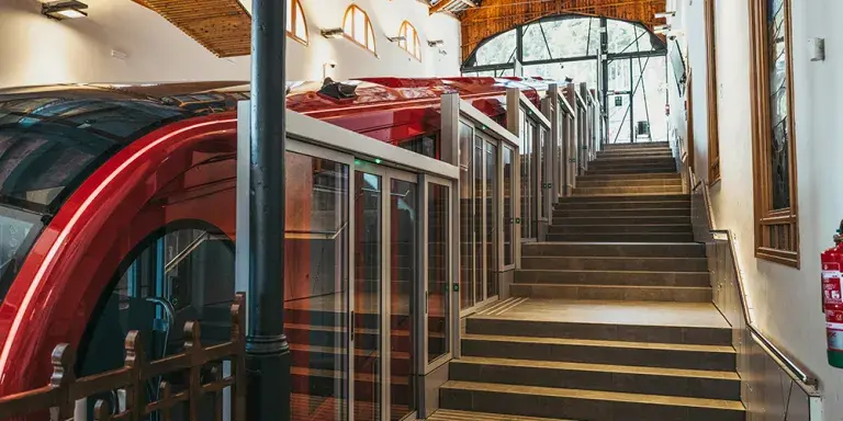 Interior de una estación con un funicular rojo moderno y puertas automáticas de vidrio a lo largo del andén con escaleras lateral.